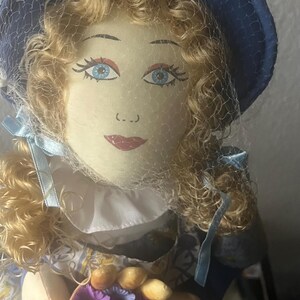 May include: A handmade doll with blonde curly hair, wearing a blue hat with a net veil, a white collar, and a blue and white dress. The doll is holding a basket of flowers and bread.