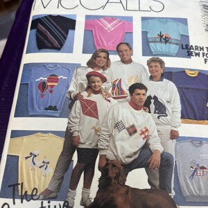 May include: A vintage McCall's sewing pattern for sweatshirts. The cover features a family wearing various sweatshirts with designs like sailboats, hot air balloons, and geometric patterns. A golden retriever dog is in the foreground. The text reads "The Creative Sweatshirt".