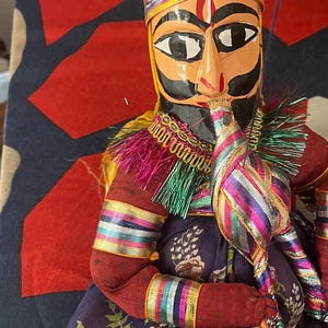 May include: A handcrafted wooden puppet with a painted face, wearing an orange turban and a colorful costume. The puppet has a black mustache and beard, and is adorned with gold and rainbow accents. The puppet is holding a decorative item.