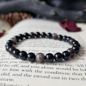 Könnte beinhalten: Ein Armband mit schwarzen und graubraunen runden Perlen, das auf einem offenen Buch liegt. Die Perlen haben eine glänzende Oberfläche und sind auf einem elastischen Band aufgefädelt. Das Buch hat Text auf den Seiten.