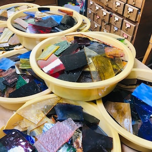 May include: Several yellow plastic tubs filled with colorful stained glass pieces. The glass shards are various shapes and sizes, with colors including blue, red, green, and yellow. The tubs are arranged on a light-colored surface.