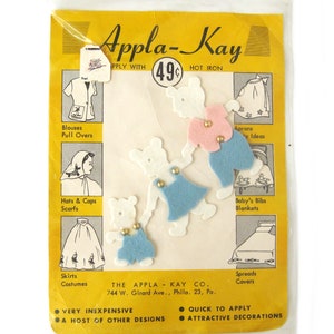Op de afbeelding: Een pakket Appla-Kay strijkbare vilt-applicaties met vier witte viltberen met verschillende gekleurde viltkleding. Het pakket is geel met zwarte tekst en afbeeldingen van suggesties voor het gebruik van de applicaties, waaronder blouses, truien, hoeden, mutsen, sjaals, rokken, kostuums, schorten, baby-slabbetjes, dekens en spreien. Het pakket bevat ook de tekst "Strijken met heet strijkijzer" en "49¢".
