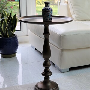 May include: A bronze side table with a circular top and ornate base. A small, blue and white patterned vase is on the table. The table is near a white sofa and a potted plant. The table is approximately 76 cm tall.