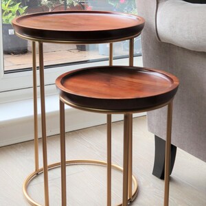 May include: Two round wooden nesting tables with gold metal frames. The larger table is on top of the smaller table.