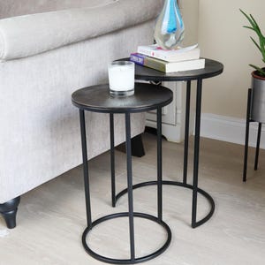 May include: A set of two black metal nesting side tables with round, textured tops. The top table has a white candle on it. The bottom table has a stack of books and a blue glass sculpture on top.