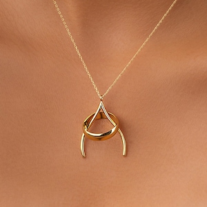 May include: A gold-colored pendant necklace featuring a unique design. The pendant has a teardrop shape at the top, a circular ring in the middle, and two curved lines extending downwards. The necklace chain is also gold.