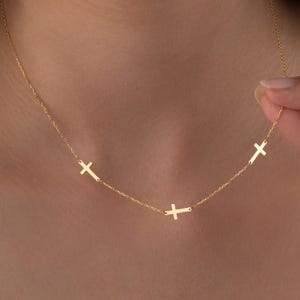May include: A delicate gold necklace featuring three small cross pendants. The crosses are evenly spaced along a fine chain, creating a simple yet elegant design. The necklace is shown against a neutral background, highlighting its minimalist style.