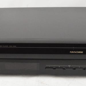 May include: A black Yamaha Natural Sound Compact Disc Player CDC-505 featuring a PlayXchange tray, a power button, a digital display area, and multiple control buttons on the front panel. The device has a rectangular shape with a matte finish.