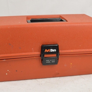 May include: A rectangular orange plastic storage box with a black handle and a black latch. The latch features the text ArtBin, Made in U.S.A., and Model 8413. The exterior surface has a textured finish with minor signs of wear and small paint spots.