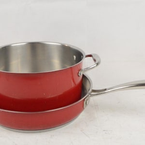 Puede incluir: Una olla roja con dos asas laterales apilada dentro de una sartén roja con un mango largo de acero inoxidable. Ambas piezas de cocina presentan un acabado exterior en rojo brillante y una superficie interior de acero pulido.
