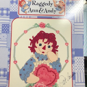 May include: Cross stitch pattern featuring Raggedy Ann holding a heart that says "I Love You" with roses and a signature by Pat Carson. The pattern is titled "I Love You" and is a Classic Raggedy Ann & Andy design.