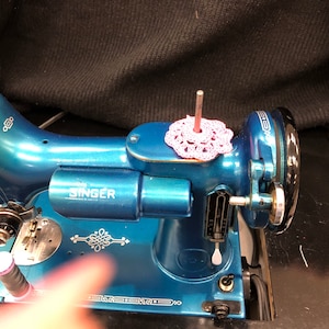 Puede incluir: Una máquina de coser Singer azul con un diseño blanco y plateado. La máquina tiene un encaje de ganchillo rosa en la parte superior de la aguja.