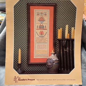 May include: A framed counted thread sampler with a cross-stitch design featuring a bowl of fruit, a house, and a flower. The sampler is in a wooden frame and is displayed with candles and a decorative vase. The text "The Counted Thread Sampler" is at the top.