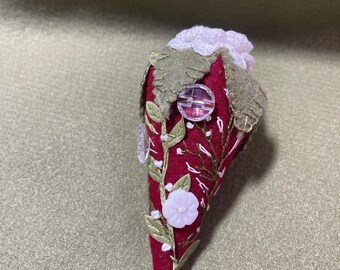 May include: A cone-shaped ornament made of deep red felt, featuring embroidered green vines and leaves, small white stitched accents, a light pink flower-shaped button, a clear circular button, and a pale pink crocheted top.