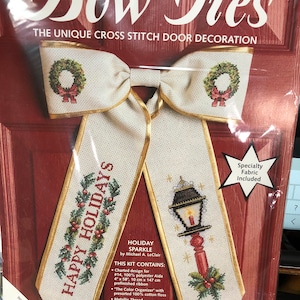 Puede incluir: Un kit de punto de cruz para una decoración de puerta con lazo de Navidad. El kit incluye una cinta preacabada, hilo de algodón 100%, hilo metálico, una aguja, tela para una cinta y las instrucciones. El diseño presenta una corona y una farola con las palabras "Felices Fiestas" en la cinta.