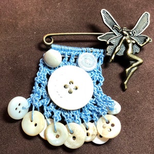 Puede incluir: Un broche de crochet azul con un dije de hada de metal y botones blancos. El broche tiene un cierre de pasador de seguridad.