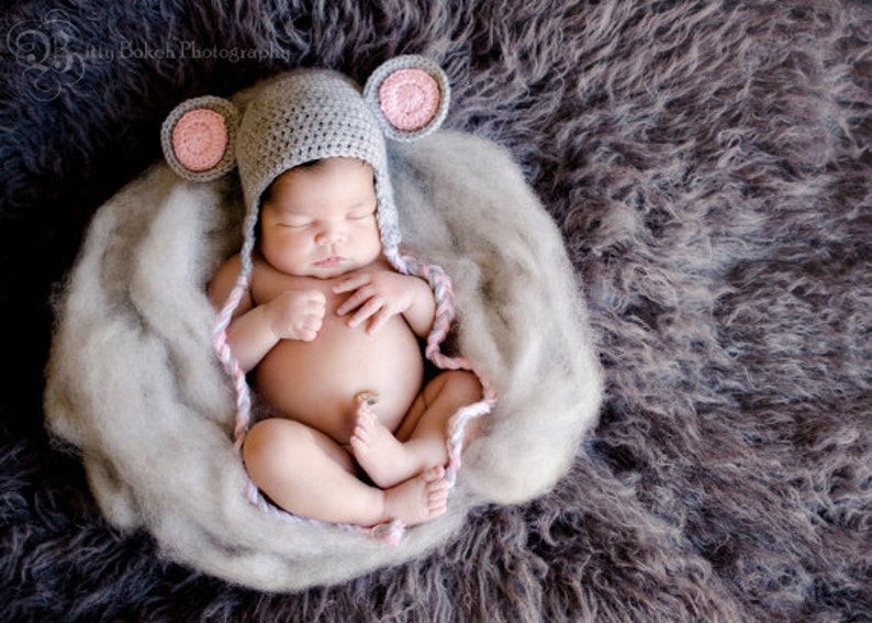 K&ouml;nnte beinhalten: Ein neugeborenes Baby tr&auml;gt eine graue geh&auml;kelte M&uuml;tzen mit rosa Ohren und schl&auml;ft auf einer flauschigen grauen Decke.