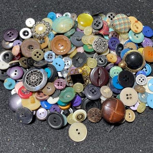 May include: A collection of various buttons in different sizes, shapes, and colors. The buttons include a range of materials, such as plastic, metal, and fabric. Colors include blue, brown, black, white, yellow, and purple. Some buttons have decorative elements.