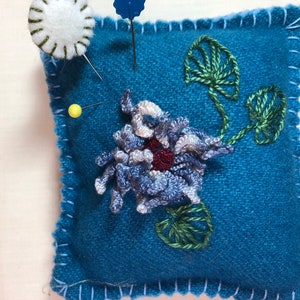 May include: A teal square pincushion with a white, hand-stitched border. It features an embroidered blue and white flower with a maroon centre and green leaves. Several pins are inserted into the cushion.