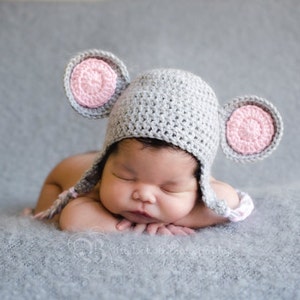 K&ouml;nnte beinhalten: Ein neugeborenes Baby tr&auml;gt eine graue H&auml;kelm&uuml;tze mit rosa Ohren und schl&auml;ft auf einer grauen, flauschigen Decke.