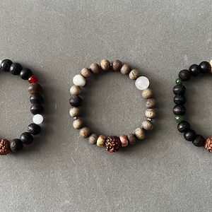 Könnte beinhalten: Drei Holzperlenarmbänder mit unterschiedlich farbigen Perlen und Anhängern. Die Armbänder haben eine braune, schwarze und schwarz-grüne Farbgebung. Jedes Armband hat einen anderen Anhänger, darunter einen braunen, einen weißen und einen gelben Anhänger.
