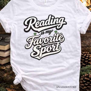 Peut inclure: T-shirt blanc avec l'inscription "Reading is my Favorite Sport" en noir et blanc. Le t-shirt a un col rond et des manches courtes. Le texte comprend deux petits cœurs verts. Le t-shirt est noué à la taille.
