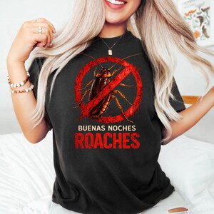 May include: Black t-shirt with a graphic design featuring a cockroach inside a red "no" symbol. The text "BUENAS NOCHES ROACHES" is printed below the graphic in red letters. The shirt is a dark color.