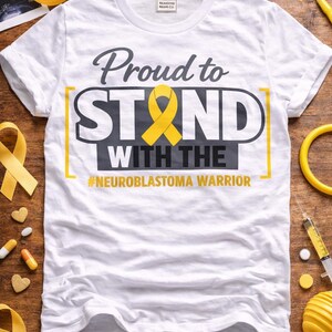 Peut inclure: T-shirt blanc avec l'inscription "Proud to STAND with the #NEUROBLASTOMA WARRIOR" en gris et jaune. Un ruban jaune est au centre du motif. Le t-shirt est entouré de rubans jaunes, de fournitures médicales et de bonbons en forme de cœur.