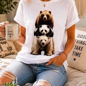 Peut inclure: Un t-shirt blanc à manches courtes avec une impression graphique représentant trois ours empilés : un grizzly brun en haut, un panda noir et blanc au milieu et un ours polaire brun clair en bas, tous debout en colonne verticale.