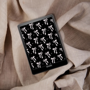 Puede incluir: Un lector electrónico Kindle negro que muestra un patrón de lazos blancos sobre un fondo negro. El lector está sobre una tela beige. La palabra "kindle" es visible en la parte inferior.