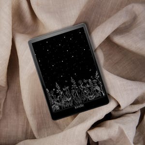 May include: A black Kindle e-reader with a screen displaying a black background with white stars and a white floral design at the bottom. The e-reader is resting on a beige fabric.