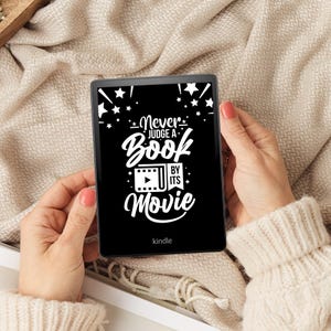 May include: A black and white Kindle e-reader with a quote that reads "Never judge a book by its movie" on the screen. The e-reader is being held by a person's hands.