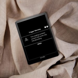 May include: A black Kindle e-reader displaying a "Trigger Warning" message on its screen. The warning includes an exclamation point symbol and the text "This reading contains content that some may find disturbing." The e-reader is resting on a beige fabric.