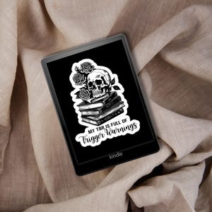 Puede incluir: Ilustración en blanco y negro de una calavera con rosas y libros, con el texto "My TBR is full of trigger warnings" en un lector electrónico Kindle negro.