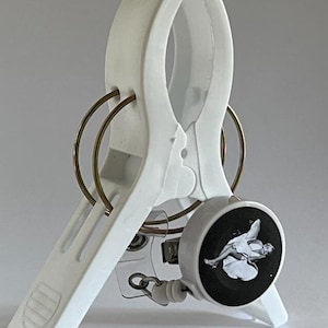 Op de afbeelding: Een witte plastic badgehouder met een oprolmechanisme. De rol heeft een zwart-wit afbeelding van een vrouw. De badgehouder heeft twee goudkleurige ringen.
