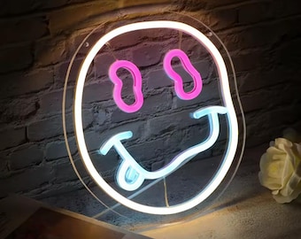 May include: A neon sign in the shape of a smiling face with a tongue sticking out. The eyes are pink, the mouth and tongue are light blue, and the outline is white. The sign is mounted on a clear acrylic backing.