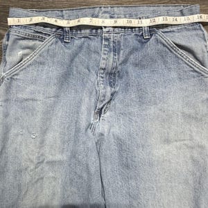 May include: Light blue denim work pants with a measuring tape across the waist. The tape reads approximately 16 inches across. The pants have front pockets and a button and zipper closure. The denim has some wear and tear.