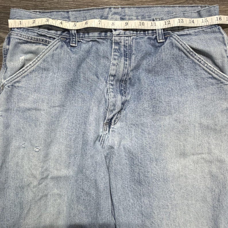 May include: Light blue denim work pants with a measuring tape across the waist. The tape reads approximately 16 inches across. The pants have front pockets and a button and zipper closure. The denim has some wear and tear.