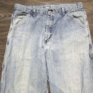 May include: Light blue denim work pants with multiple pockets and a button closure. The pants show signs of wear and tear, with some fading and small holes. The jeans have a classic, relaxed fit.