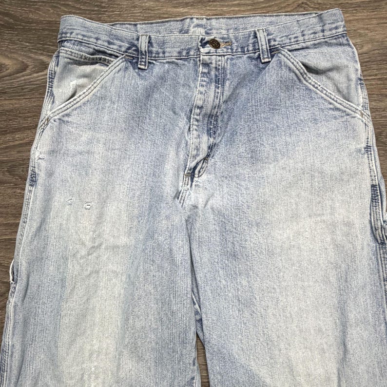 May include: Light blue denim work pants with multiple pockets and a button closure. The pants show signs of wear and tear, with some fading and small holes. The jeans have a classic, relaxed fit.