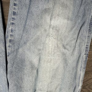 May include: Close-up of light blue denim jeans with visible wear and tear. The fabric shows faded areas and subtle variations in color, suggesting regular use. The jeans have a classic five-pocket design.