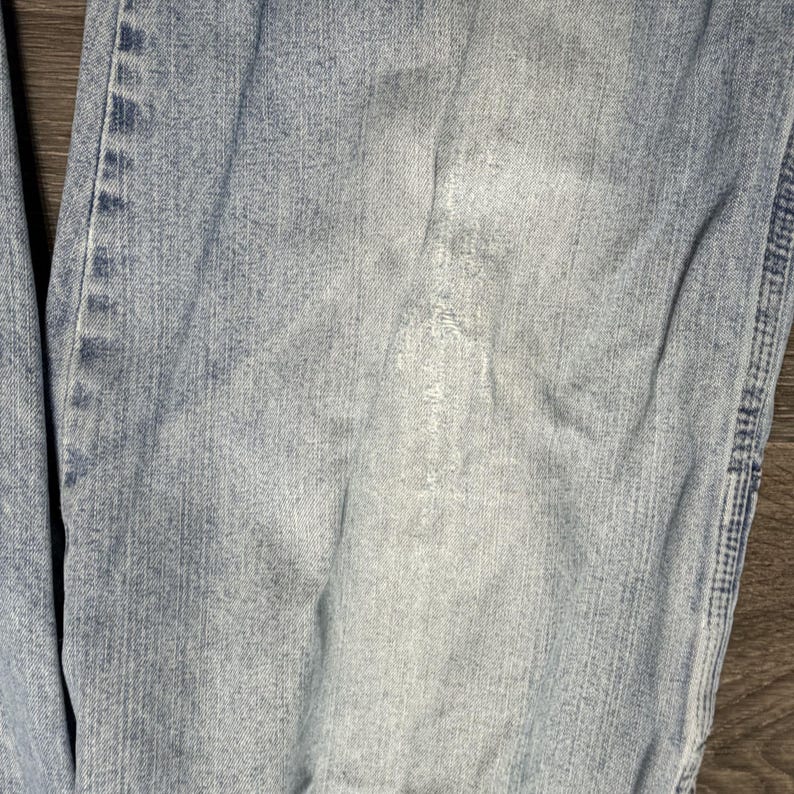 May include: Close-up of light blue denim jeans with visible wear and tear. The fabric shows faded areas and subtle variations in color, suggesting regular use. The jeans have a classic five-pocket design.