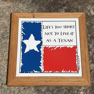 May include: A framed tile with the Texas flag design. It features a white star on a blue background, a red rectangle, and the text "LIFE'S TOO SHORT NOT TO LIVE IT AS A TEXAN". The frame is made of light brown wood.