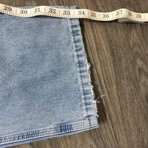 May include: Close-up of light blue denim jeans with a measuring tape showing a waist measurement. The tape reads from 29 to 39 inches. The jeans have a frayed edge.