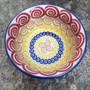 May include: A round ceramic bowl featuring a hand-painted pattern of concentric circles and spirals. The design includes a wide outer band of red spirals, followed by rings of tan, yellow, and a central circle of blue spirals with a tan center.