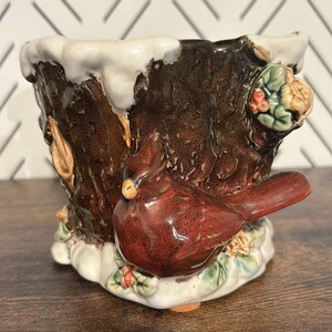 May include: A ceramic planter featuring a dark brown tree trunk design, with white detailing at the top. A red cardinal bird is perched on the side, with green leaves and red berries. The planter has a rustic, handcrafted appearance.
