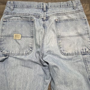 May include: Light blue denim work pants with multiple pockets and a small rectangular label on the back pocket. The pants have a faded appearance and visible stitching details.