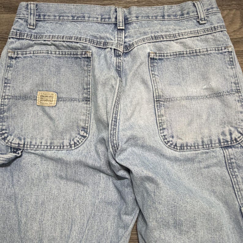 May include: Light blue denim work pants with multiple pockets and a small rectangular label on the back pocket. The pants have a faded appearance and visible stitching details.