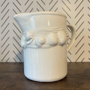 May include: A white ceramic pitcher with a handle and spout. The pitcher is decorated with a band of seashell relief around the middle. The pitcher is sitting on a dark brown surface.