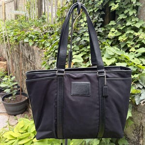 May include: Black tote bag with leather accents and handles. The bag features a rectangular leather patch with the brand name "COACH" embossed on the front. The bag has a rectangular shape with a top opening and two side pockets.